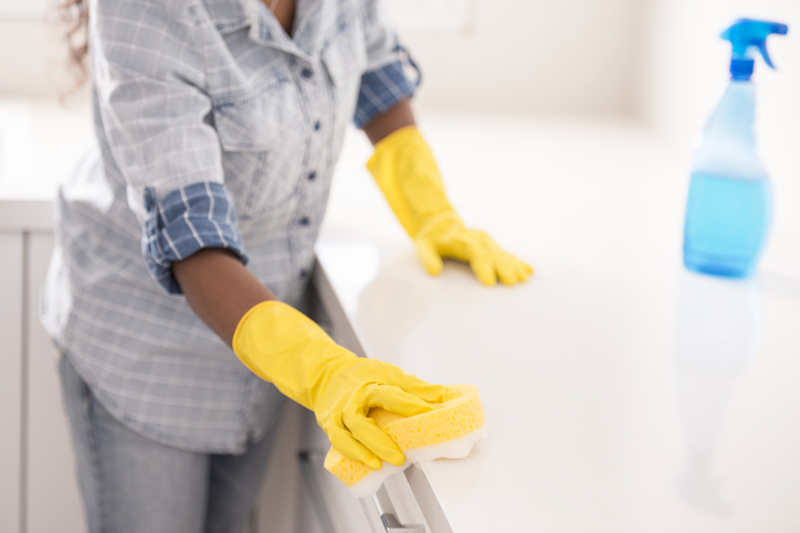 Image of a kitchen counter top being cleaned. Person wearing yellow rubber gloves holding a yellow sponge with blue leaning solution in spray bottle 